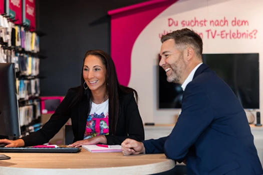 Zwei Personen beraten sich lächelnd in einem modernen Telekom-Shop am Computer.