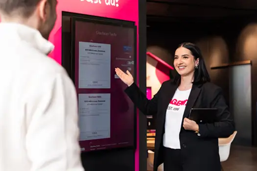 Mitarbeiterin erklärt einem Kunden an einem digitalen Infoterminal verschiedene Tarifoptionen.