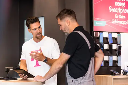 Ein Berater erklärt einem Kunden im Telekom-Shop verschiedene Smartphones am Verkaufstresen.